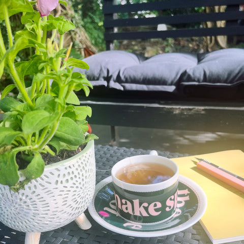 Teacup with 'Make Spot' text, plant, and notebook on a table outdoors.